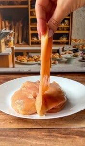 A hand lifts a raw, thin slice of chicken breast from a white plate with more raw chicken, set on a wooden table in front of a bakery-themed background with bread and pastries.