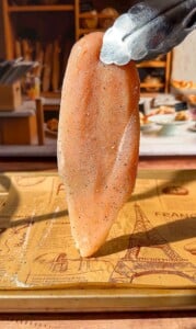 A pair of metal tongs holds up a raw, seasoned chicken breast over a wooden tray lined with parchment paper printed with Paris-themed designs.