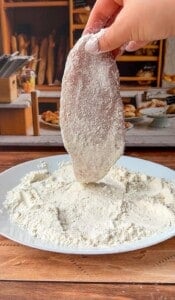 A hand dips a raw chicken breast fillet into a plate of flour on a wooden table, preparing it for cooking. In the background, there are breads and other kitchen items.