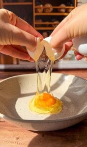 Close-up of hands cracking an egg over a beige bowl, with the egg white and yolk falling into the bowl. Sunlight highlights the scene, and shelves with items are blurred in the background.