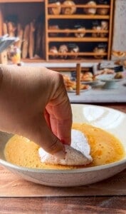 A hand dips a powdered, crescent-shaped pastry into a bowl of beaten eggs, with shelves of bread and pastries blurred in the background.
