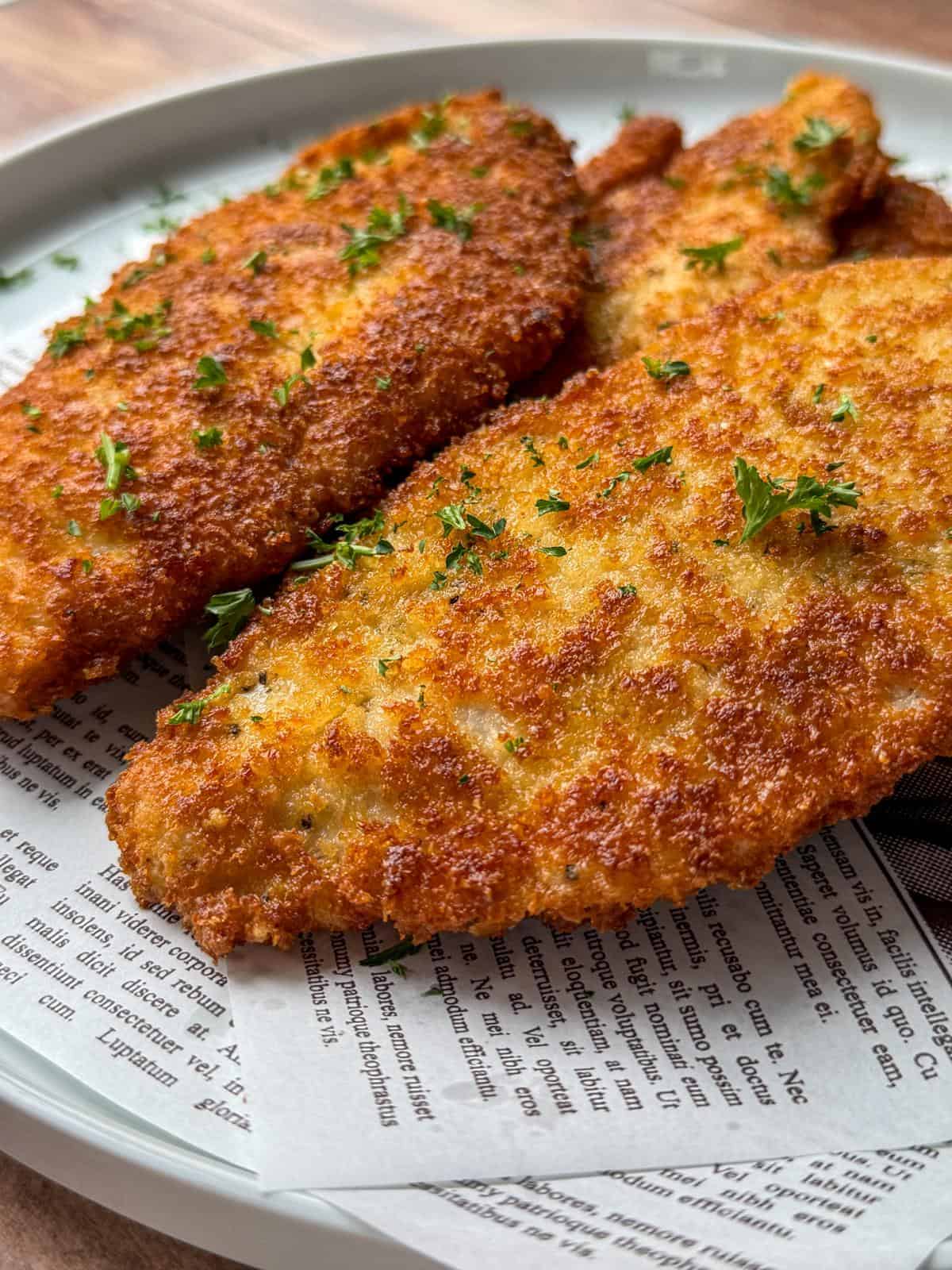 Two golden-brown breaded chicken cutlets garnished with chopped parsley, served on a plate lined with decorative newspaper-style parchment paper.
