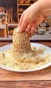 A hand dips a cone-shaped pastry into a plate of breadcrumbs, with a bakery display of bread and pastries in the background.