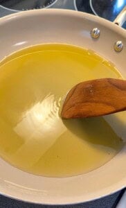 A wooden spatula stirs oil in a light-colored frying pan on a stovetop, with light reflecting off the surface of the oil.