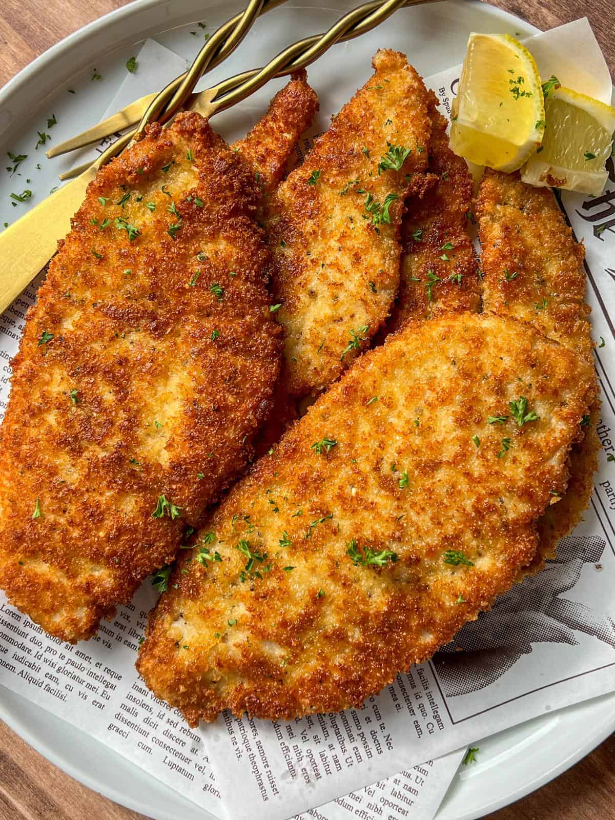 Golden, crispy breaded cutlets garnished with chopped parsley are arranged on a plate lined with newspaper print. Lemon wedges and a wooden-handled fork are placed on the side.