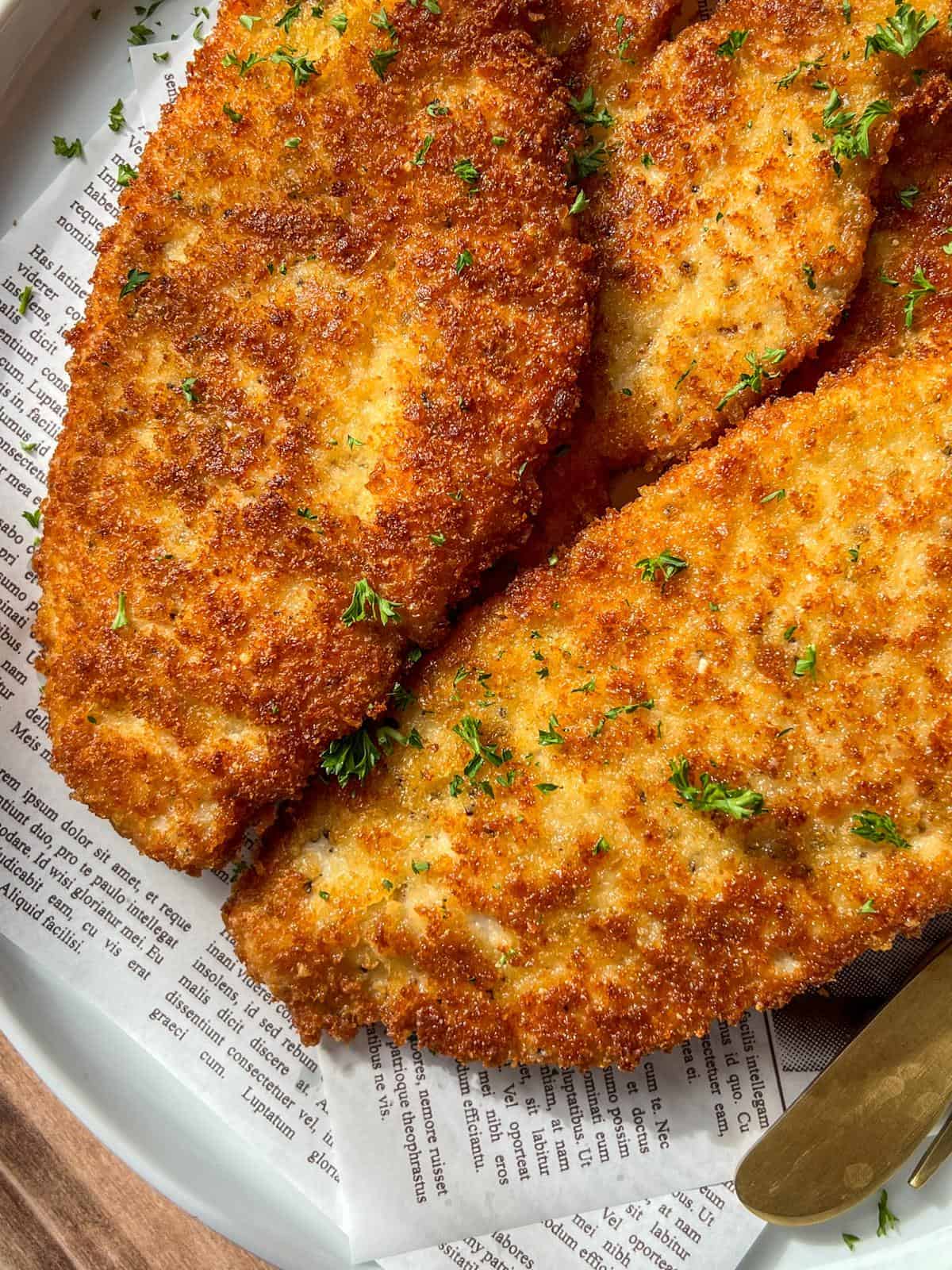 Golden, crispy breaded chicken cutlets garnished with chopped parsley are served on a plate lined with printed newspaper-style parchment paper. A knife is partially visible at the edge of the plate.