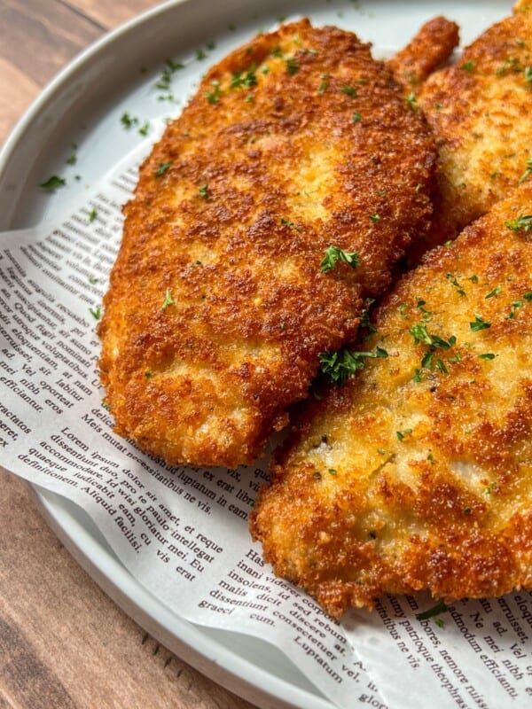 Close-up of two golden brown breaded chicken cutlets garnished with chopped parsley, served on a plate lined with decorative newspaper-print paper.