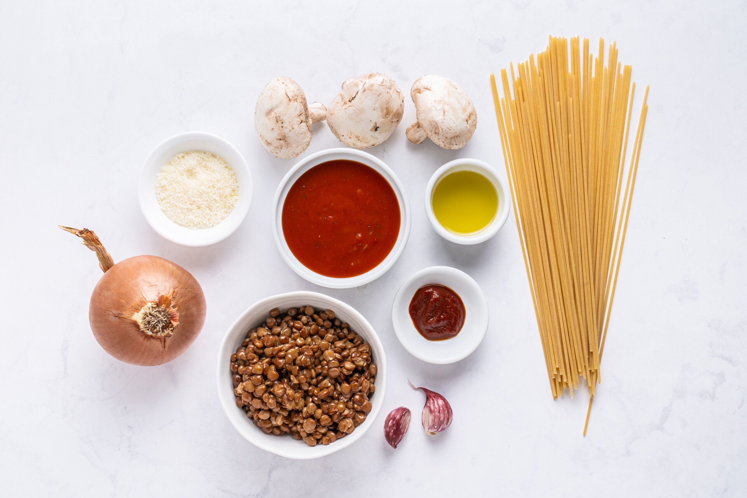 Ingredients for a pasta dish arranged on a white surface: uncooked spaghetti, whole mushrooms, yellow onion, lentils, tomato sauce, olive oil, tomato paste, grated cheese, and two garlic cloves in small bowls.
