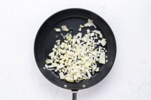Chopped onions being sautéed in a black frying pan on a white surface.
