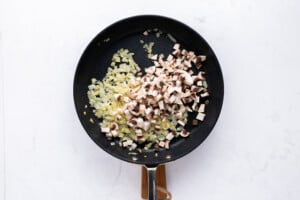 A black frying pan on a white surface contains sautéed chopped onions and freshly added chopped mushrooms, with both ingredients partially mixed.