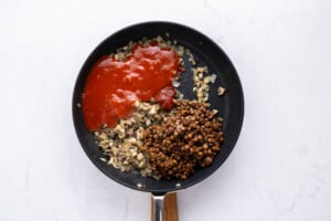 A frying pan on a white surface contains chopped onions and mushrooms, a portion of tomato sauce, and a serving of cooked lentils, all separated into sections and not yet mixed together.