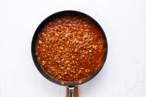 A black pan filled with a chunky red tomato sauce containing corn, lentils, and other visible vegetables, set against a white background.