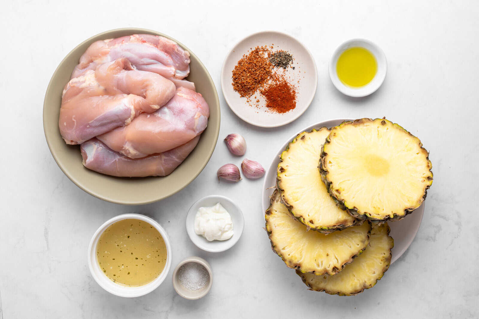 Flat lay of raw chicken thighs, sliced fresh pineapple, garlic cloves, assorted spices, olive oil, a bowl of marinade, a small dish of mayonnaise, and a dish of salt on a white surface. - 4