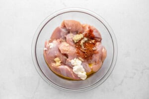 Raw chicken pieces in a glass bowl with dollops of yogurt, minced garlic, oil, and red spices on top, ready to be mixed, on a white marble surface.