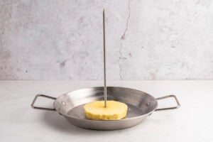A thick slice of pineapple is pierced vertically by a long metal skewer, standing upright in the center of a round metal pan with two handles, placed on a light-colored surface.