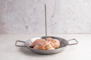 Raw, marinated chicken pieces are placed in a round metal pan with handles, and a vertical metal skewer stands upright through the center of the chicken. The background is a light, textured surface.