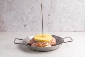 A metal pan holds raw marinated chicken pieces stacked with a pineapple slice on top, all skewered vertically with a metal rod, against a light background.