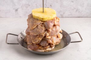 Raw marinated meat stacked on a vertical skewer with a slice of pineapple on top, placed in a metal pan with handles, against a light textured background.