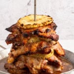 A stack of grilled, marinated meat slices layered with grilled pineapple rings, skewered vertically on a metal rod, sits on a metal pan with juices at the bottom against a light background.