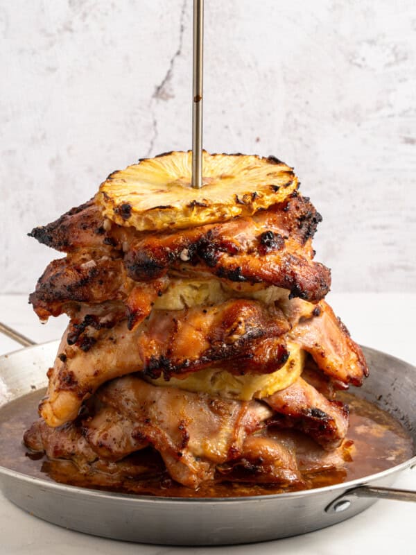 A stack of grilled, marinated meat slices layered with grilled pineapple rings, skewered vertically on a metal rod, sits on a metal pan with juices at the bottom against a light background.
