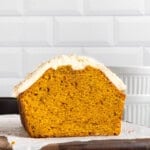 A loaf of pumpkin bread with creamy white frosting on top sits on parchment paper over a wooden board, shown cut in half to display its moist orange interior, against a white tiled background.