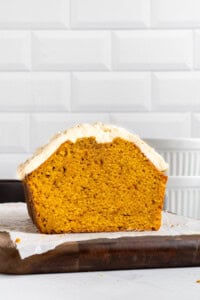 A loaf of pumpkin bread with creamy white frosting on top sits on parchment paper over a wooden board, shown cut in half to display its moist orange interior, against a white tiled background.