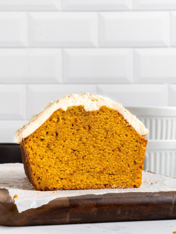 A loaf of pumpkin bread with creamy white frosting on top sits on parchment paper over a wooden board, shown cut in half to display its moist orange interior, against a white tiled background.