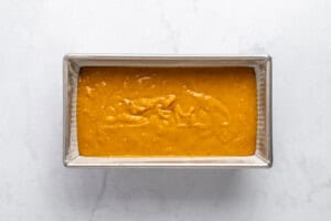 A rectangular metal baking pan filled with smooth, orange-yellow batter sits on a white marble surface, ready to be baked.