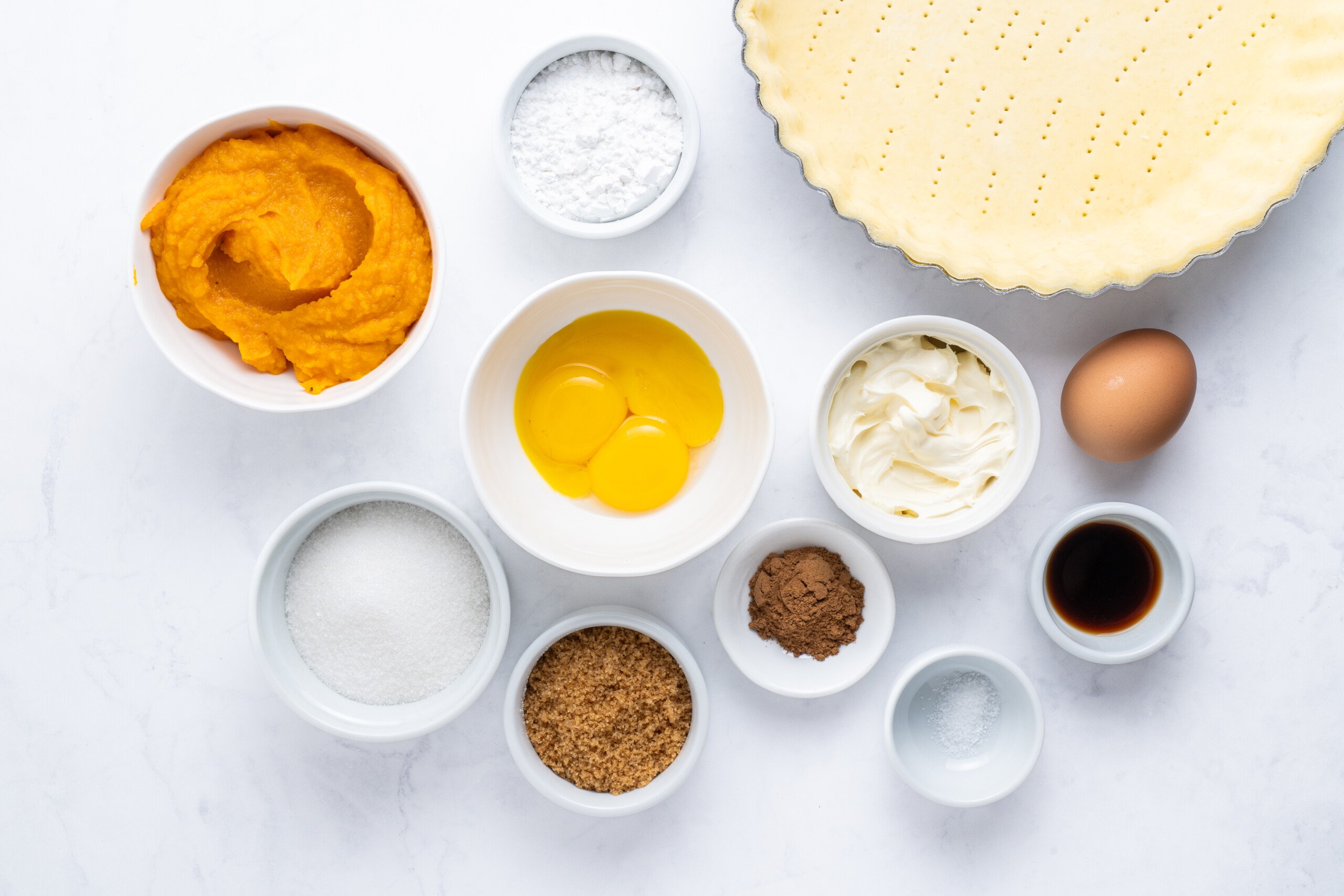 A flat lay of pumpkin pie ingredients: pumpkin purée, eggs, sugar, brown sugar, cocoa powder, salt, vanilla extract, cream cheese, powdered sugar, a whole egg, and an unbaked pie crust in a tin.
