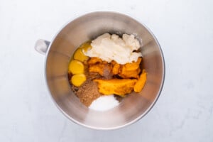 A metal mixing bowl containing eggs, pumpkin puree, sugar, spices, and a dollop of cream cheese or sour cream, set on a light-colored surface.