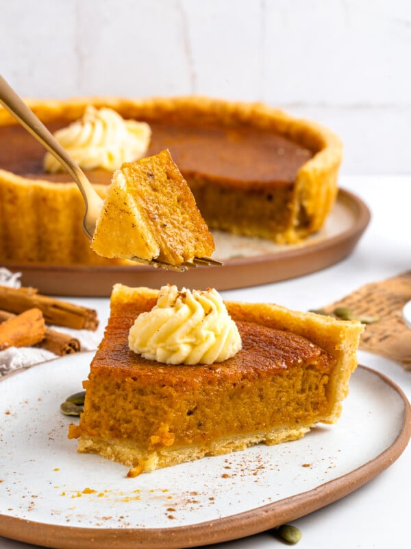 A slice of pumpkin pie with whipped cream sits on a plate, while a fork holds up a bite. The rest of the pie is in the background, along with cinnamon sticks and a light, rustic setting.
