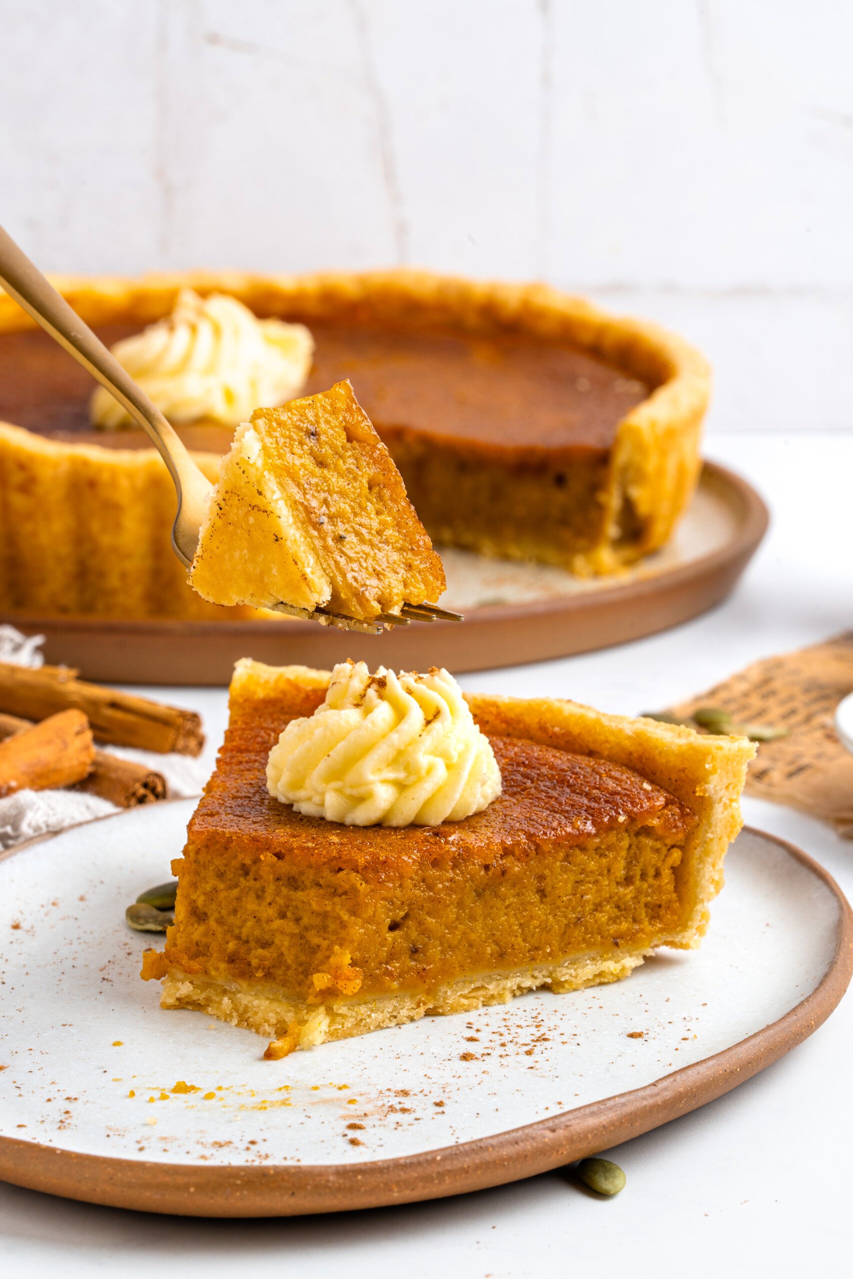 A slice of pumpkin pie with whipped cream sits on a plate, while a fork holds up a bite. The rest of the pie is in the background, along with cinnamon sticks and a light, rustic setting.