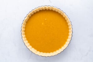 A round tart pan filled with an unbaked pumpkin pie mixture sits on a light, marble surface. The smooth orange filling is surrounded by a crimped pie crust.