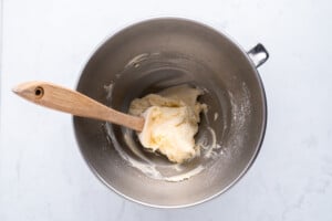 A metal mixing bowl containing creamy dough and a wooden spatula, set on a white marble surface.
