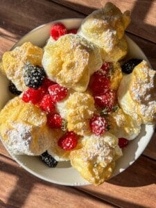 A bowl filled with cream puffs dusted with powdered sugar, garnished with fresh raspberries and blackberries, sits on a sunlit wooden surface—perfect alongside viral high protein mini pancakes for a delightful breakfast spread.
