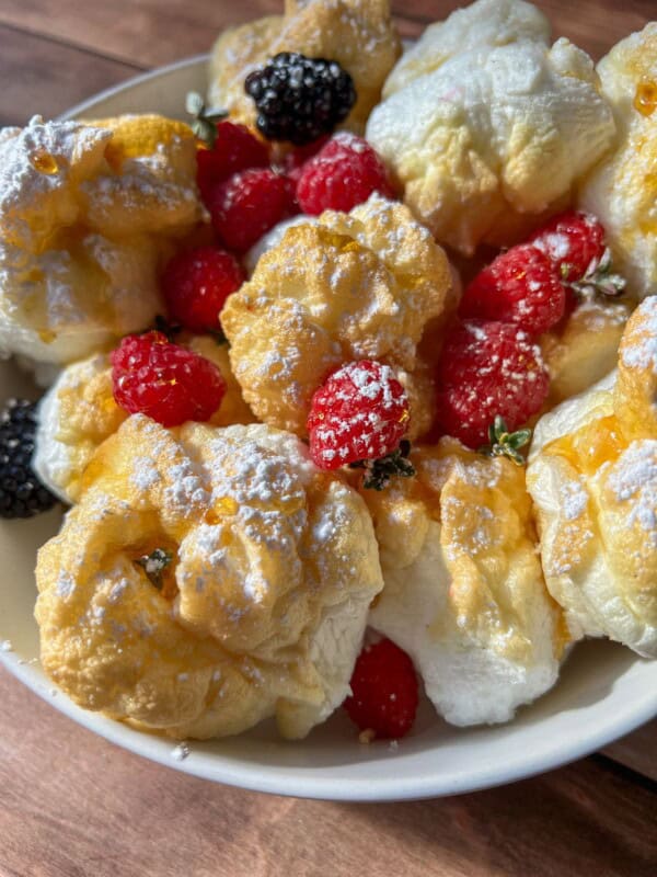 A bowl of fluffy cream puffs dusted with powdered sugar, garnished with fresh raspberries and blackberries, sits beside High Protein Mini Pancakes (Viral) on a rustic wooden table.