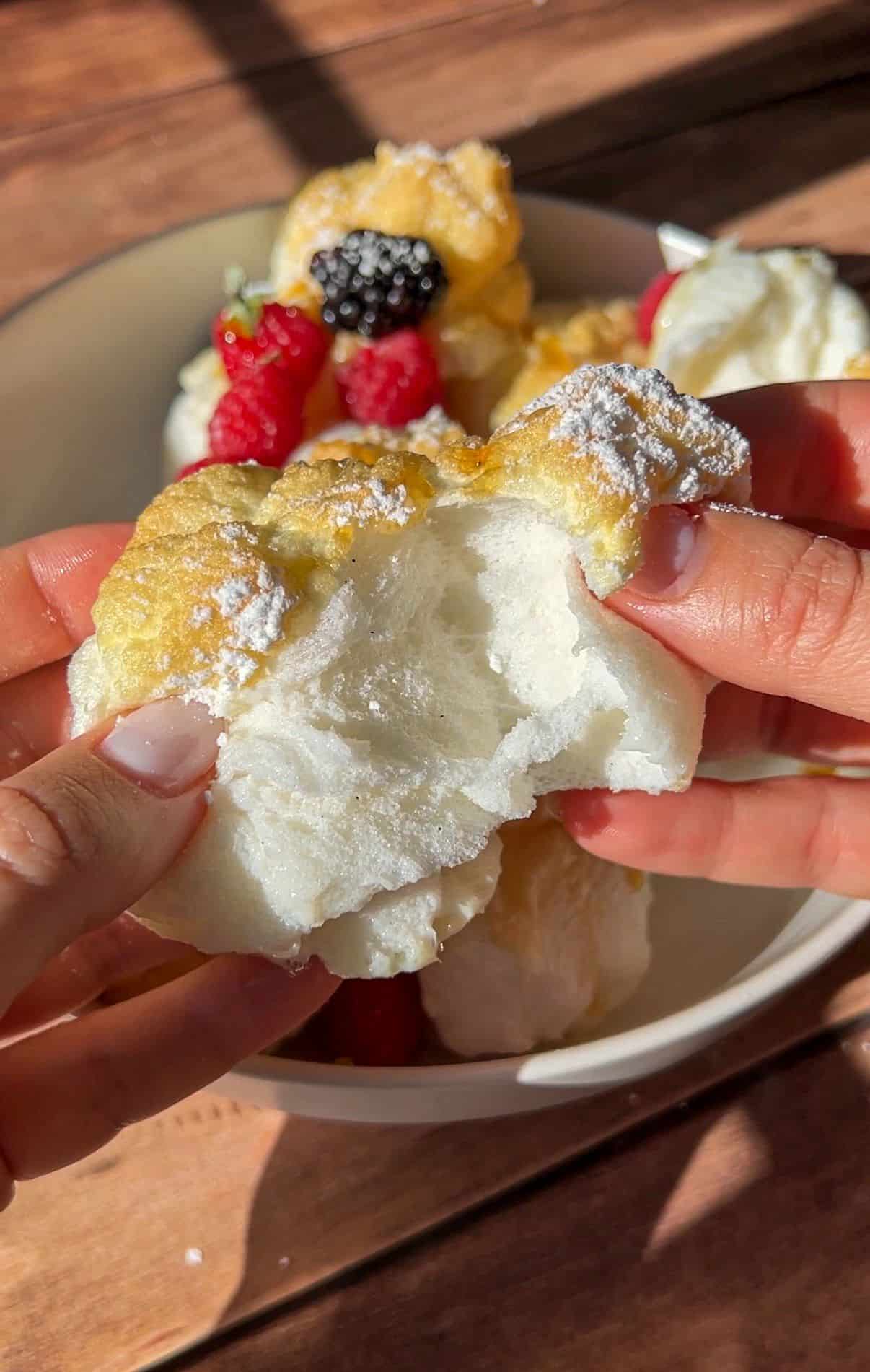 A person breaks open fluffy, golden High Protein Mini Pancakes (Viral) dusted with powdered sugar. Fresh berries are visible on other pieces of the pancakes in a white bowl on a wooden table. - 5