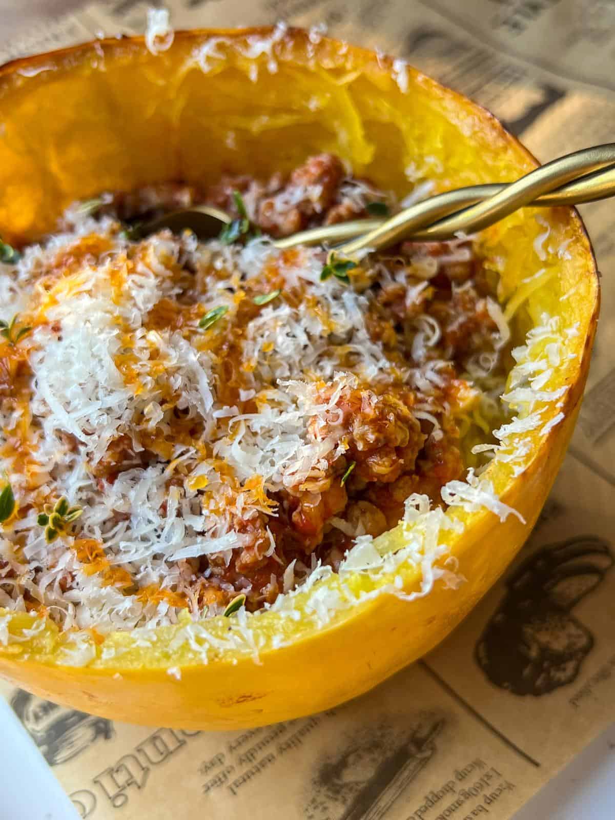 A close-up of spaghetti squash used as a bowl, filled with a tomato-based meat sauce and topped with grated cheese and herbs, with a fork resting inside. The dish sits on a newspaper-print surface.