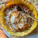 A baked spaghetti squash filled with tomato meat sauce and topped with grated cheese, served in its shell with a spoon. The dish sits on a white plate and a sheet of brown paper.