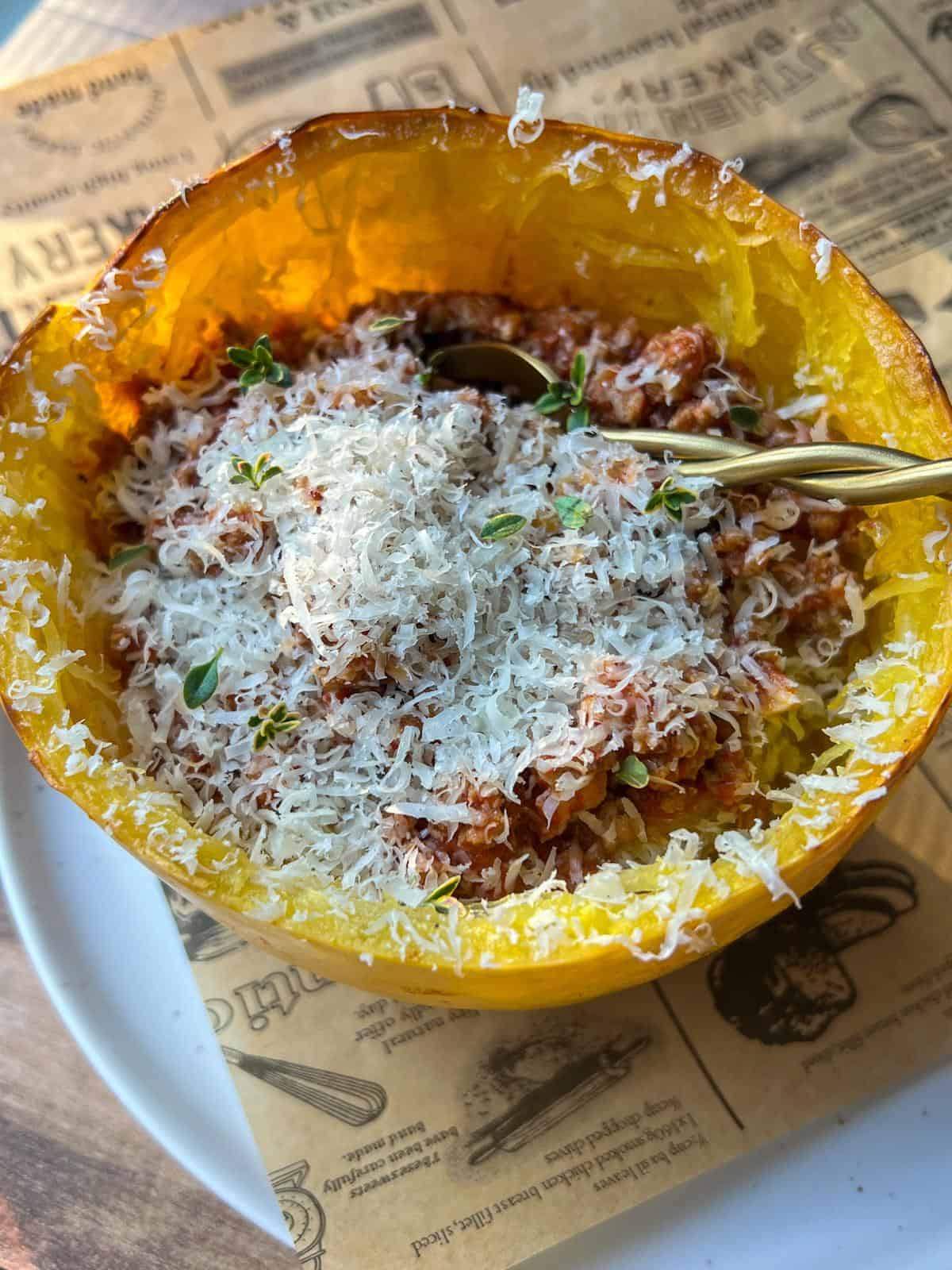 A halved spaghetti squash used as a bowl, filled with a tomato-based sauce, topped with grated Parmesan cheese and fresh herbs, with a golden spoon inside. The dish sits on a plate with parchment paper underneath.