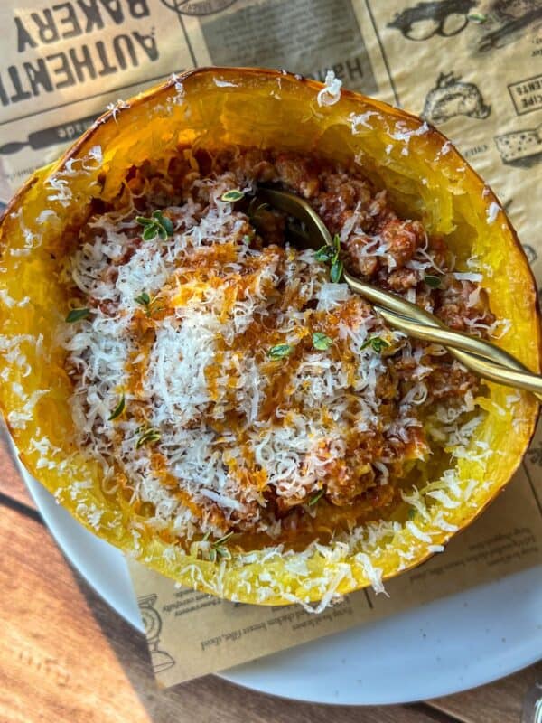 A baked spaghetti squash filled with tomato meat sauce and topped with grated cheese, served in its shell with a spoon. The dish sits on a white plate and a sheet of brown paper.