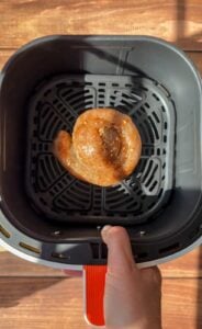 A hand is holding an air fryer basket containing a single coiled raw sausage, seasoned with spices, on a black grill tray. The background is a wooden surface illuminated by sunlight.