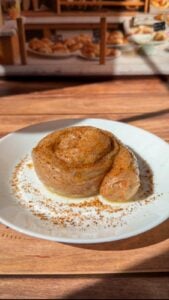 A cinnamon roll dusted with cinnamon sits on a white plate atop a wooden table, with a bakery display of various pastries blurred in the background.