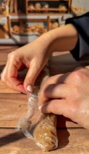 A person’s hands are unwrapping a long, cylindrical food item covered in plastic wrap on a wooden table. Shelves with baked goods are visible in the blurred background.