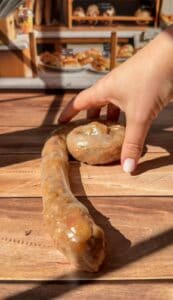 A hand with pale pink nail polish arranges a long, coiled sausage and a round sausage on a wooden table in a bakery, with shelves of bread and pastries blurred in the background.