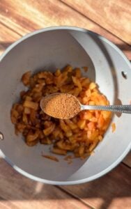A spoonful of ground spices is held above a pot containing sautéed chopped onions and vegetables, ready to be added to the mixture. Warm sunlight casts shadows on the wooden surface beneath.