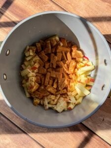 A pot containing chopped apples topped with ground cinnamon, sitting on a wooden surface with sunlight casting shadows across the scene.