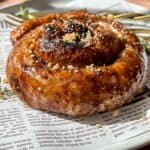 A close-up of a glazed cinnamon roll topped with crushed nuts, served on a white plate lined with newspaper print, with a fork and sprigs of thyme in the background.