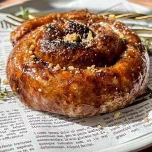 A close-up of a glazed cinnamon roll topped with crushed nuts, served on a white plate lined with newspaper print, with a fork and sprigs of thyme in the background.