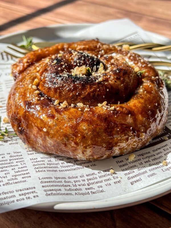 A close-up of a glazed cinnamon roll topped with crushed nuts, served on a white plate lined with newspaper print, with a fork and sprigs of thyme in the background.